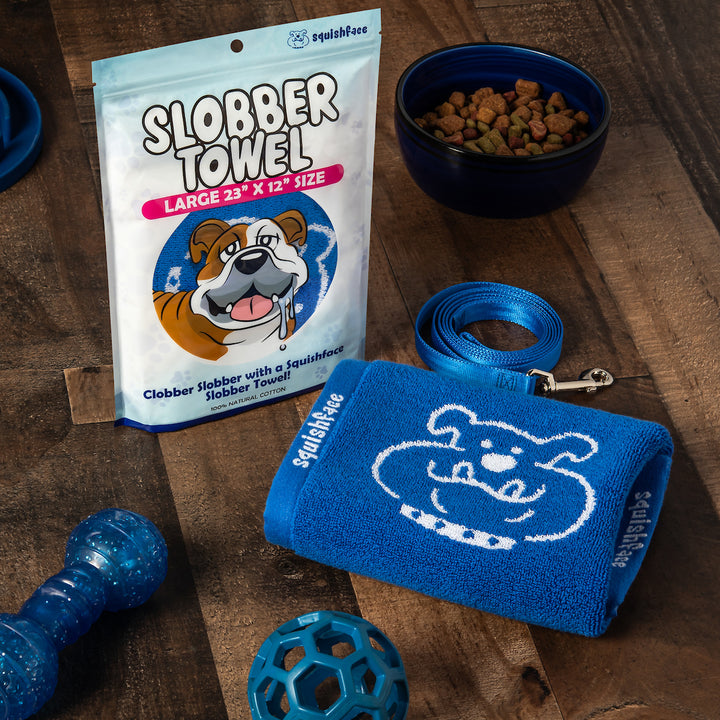 Dog accessories including a blue towel, leash, and chew toy on a wooden surface with a Slobber Towel package.